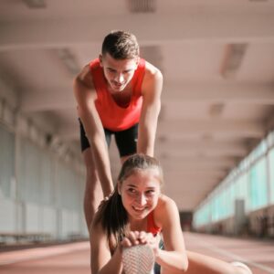 Cheerful sportswoman sitting on floor and stretching legs while man pushing back forward helping with warm up