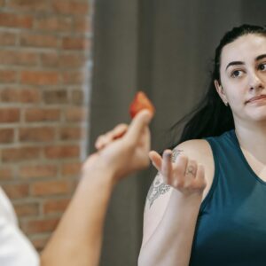 Unrecognizable personal coach offering ripe berry to ethnic overweight female in activewear while helping to lose weight in gym near brick wall