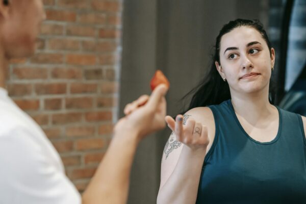 Unrecognizable personal coach offering ripe berry to ethnic overweight female in activewear while helping to lose weight in gym near brick wall